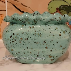 May include: A turquoise ceramic jar with a ruffled rim and a rounded body. The jar is decorated with brown speckles. The text "Kookaburra Ceramics" is written on the front. The jar is suitable for storing small items.