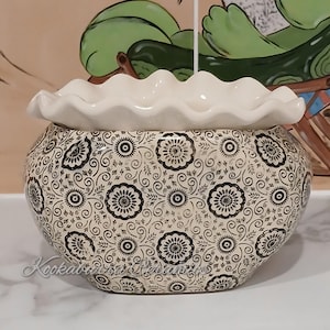 May include: A ceramic decorative pot with a cream-coloured, ruffled rim. The body of the pot is patterned with black floral and swirl designs on a cream background. The pot has a rounded shape and is likely used for decorative purposes.