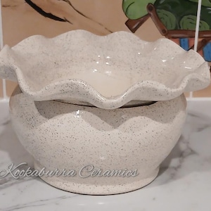 May include: A speckled, cream-coloured ceramic oil warmer with a ruffled top and a rounded base. The warmer has a speckled pattern and the text "Kookaburra Ceramics" is visible on the base.