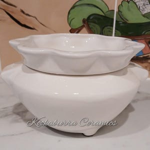 May include: A white ceramic wax warmer with a scalloped edge bowl sitting atop a rounded base. The base features a smooth, glossy finish and small feet. The text Kookaburra Ceramics is printed in a cursive script on the front of the ceramic base.