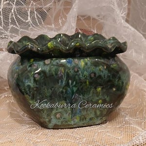 May include: A glossy ceramic planter with a ruffled, wavy rim and a mottled glaze featuring deep shades of forest green, blue, and hints of yellow. The front of the rounded vessel displays the white cursive text Kookaburra Ceramics.