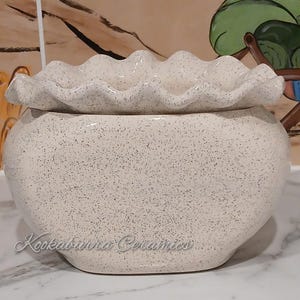 May include: A speckled, off-white ceramic container with a ruffled lid. The container has a rounded shape and a speckled pattern. The text "Kookaburra Ceramics" is written on the front of the container. The container is sitting on a white surface.
