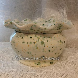 May include: A speckled ceramic container with a ruffled lid. The off-white container is decorated with green and yellow speckles. The lid has a wavy edge. The text "Kookaburra Ceramics" is written on the front of the container.