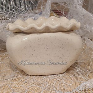 May include: A cream-coloured ceramic bowl with a speckled finish and a ruffled, wavy rim. The front of the bowl features the text Kookaburra Ceramics in a cursive font. The bowl has a rounded, bulbous body and a smooth, glossy surface texture.