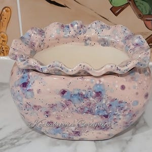 May include: A ceramic bowl with a frilled edge, in a light pink hue with blue and purple speckled detailing. The interior is a smooth, off-white colour. The text "Kookaburra Ceramics" is visible on the front.