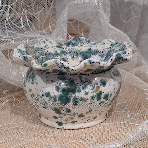 May include: A ceramic bowl with a ruffled, scalloped rim, featuring a white base with a speckled pattern of blue, teal, and gold paint. The text Kookaburra Ceramics is printed in a cursive script across the front of the rounded body.