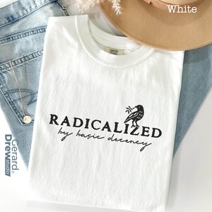 May include: White t-shirt with the text "RADICALIZED by basic decency" in black. A black bird graphic with a sprig is on the right. The shirt is on top of blue jeans, a tan hat, and a silver necklace.