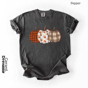 Könnte beinhalten: Ein anthrazitfarbenes Kurzarm-T-Shirt mit einer Grafik von drei Kürbissen in der Mitte. Der linke Kürbis hat ein orangefarbenes Büffelkaromuster, der mittlere ist weiß mit orangefarbenen Blumenakzenten und der rechte hat ein braunes Karomuster.