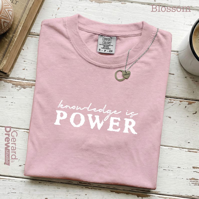 May include: A light pink t-shirt with the words "knowledge is POWER" printed in white. A silver necklace with a heart pendant rests on the shirt. The shirt is folded on a wooden surface.