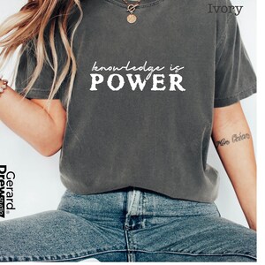 May include: A dark gray t-shirt with the words "knowledge is POWER" printed in white. The shirt is a casual style, likely made of cotton or a similar fabric. The shirt is a neutral color, suitable for various outfits.