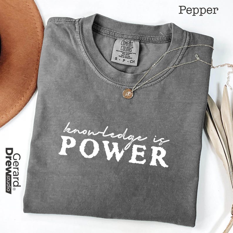 May include: A gray Comfort Colors t-shirt with the text "knowledge is POWER" in white. A gold-colored necklace with a pendant is draped over the shirt. The shirt is folded and displayed on a white surface.