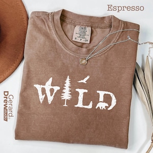 May include: Espresso-colored t-shirt with the word "WILD" in white, featuring a tree, bird, and bear graphic. A silver necklace with a pendant rests on the shirt. The shirt is folded and has a Comfort Colors tag.
