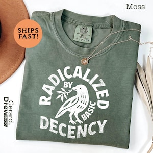 Peut inclure: T-shirt vert mousse avec l'inscription blanche "RADICALIZED BY BASIC DECENCY" et un motif d'oiseau. Un collier doré et un chapeau marron sont également visibles. Taille petite.