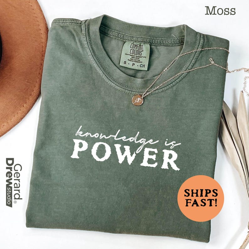 May include: A moss green t-shirt with the words "knowledge is POWER" in white. The shirt has a crew neck and short sleeves. A gold necklace with a pendant is on the shirt. The shirt is labeled "COMFORT COLORS".