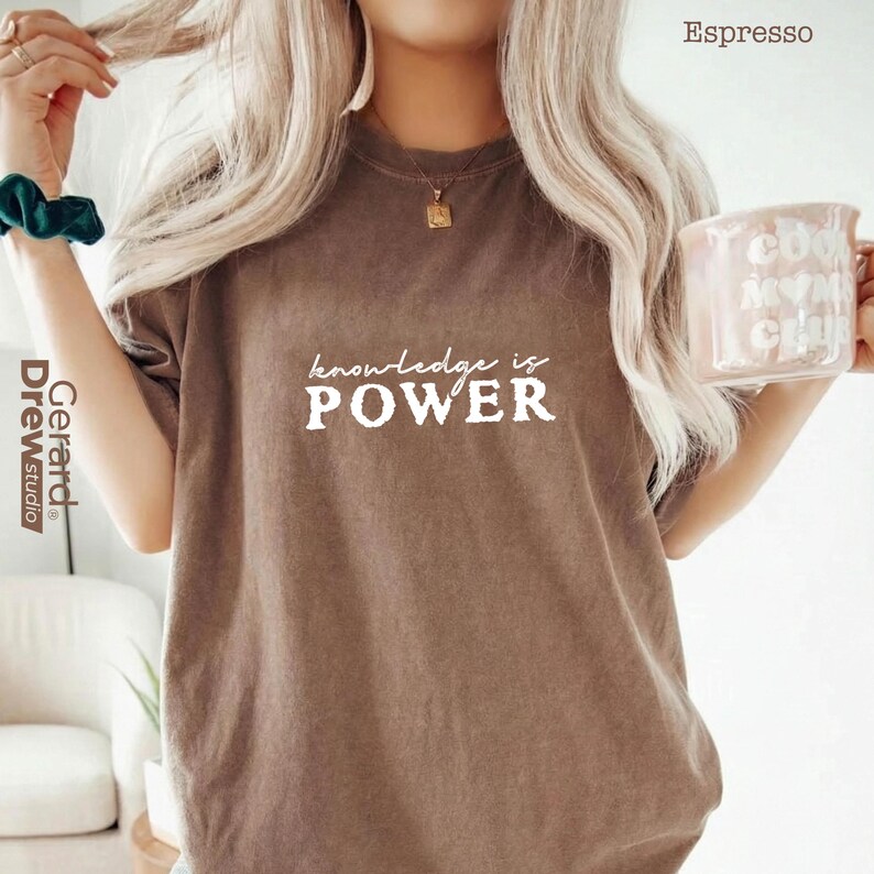 May include: Espresso-colored t-shirt with the text "knowledge is POWER" in white. The shirt is a solid color and has a crew neck. A mug with text is held in the hand.