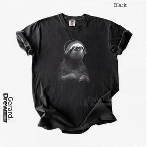 Peut inclure: Un t-shirt noir à manches courtes avec une impression graphique représentant un paresseux portant un casque audio. Le paresseux est illustré dans des tons de gris et de noir, regardant vers le haut avec une expression calme.