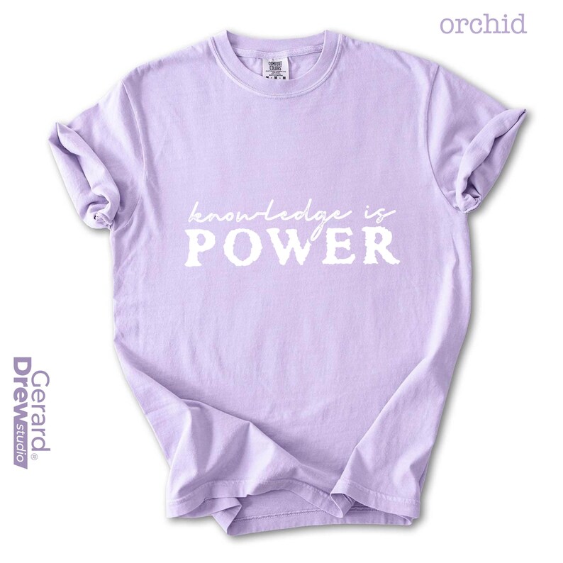 May include: Orchid-colored t-shirt with the words "knowledge is POWER" in white. The shirt has a crew neck and short sleeves. The brand name "Gerard Drew Studio" is printed on the left side.