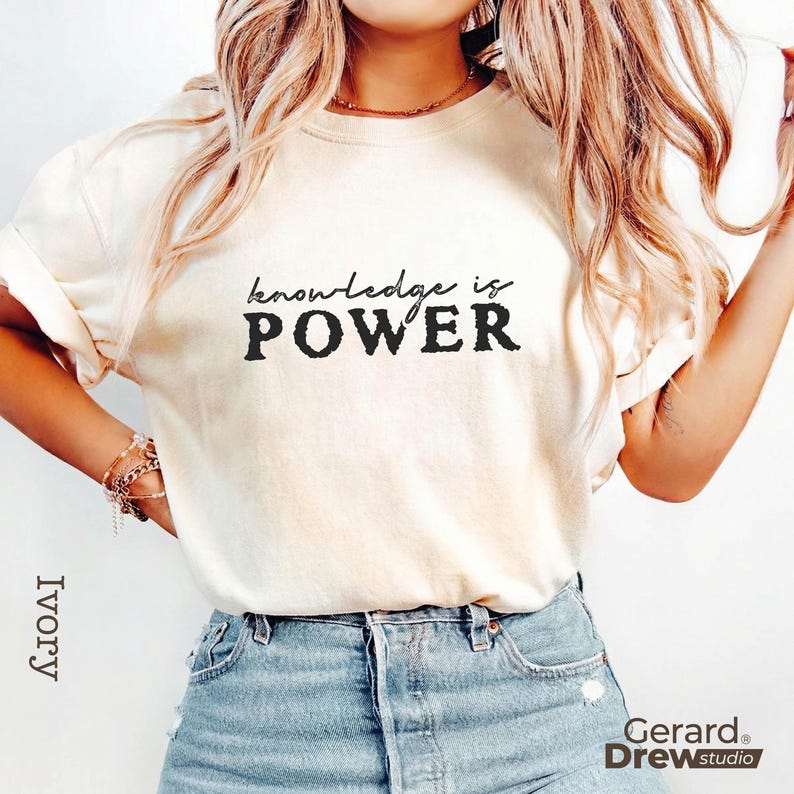May include: Ivory-colored t-shirt with the text "knowledge is POWER" in black. The shirt is paired with blue denim jeans. The brand name "Gerard Drew studio" is visible in the bottom right corner.