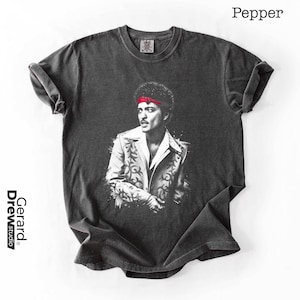 May include: A dark gray t-shirt featuring a black and white portrait of a person wearing a red headband and a patterned jacket. The text "Pepper" is at the top right, and "Gerard Drewstudio" is on the left side.