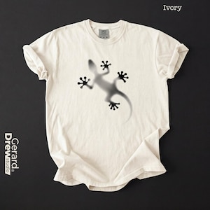 Könnte beinhalten: Ein elfenbeinfarbenes Kurzarm-T-Shirt mit einer verschwommenen, schwarzen Grafik im Airbrush-Stil, die einen Gecko in der Mitte der Brust zeigt. Das Shirt hat einen Rundhalsausschnitt und ein sichtbares Markenetikett am Innenkragen. Oben rechts steht Ivory.