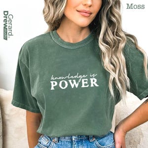 May include: Moss green t-shirt with the text "knowledge is POWER" in white. The shirt has short sleeves and a crew neck. The brand "Gerard Drew studio" is printed on the left side.