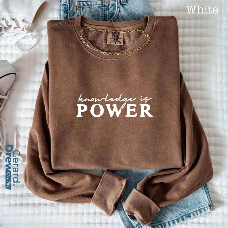 May include: A brown sweatshirt with the words "knowledge is POWER" in white lettering. The long-sleeved top is folded, with a gold chain necklace resting on it. The sweatshirt is on top of blue jeans.