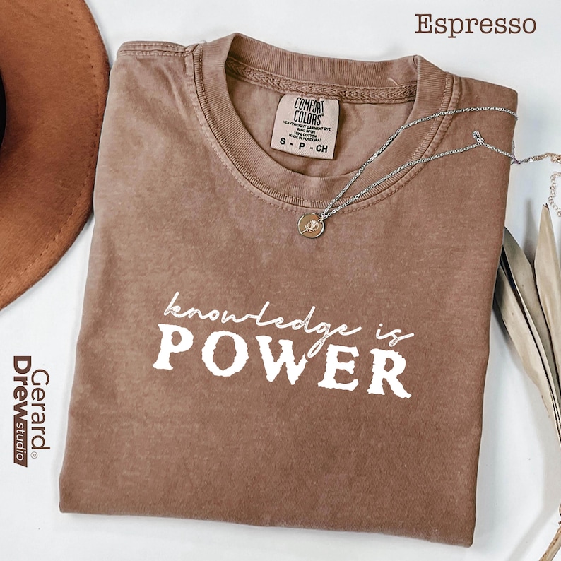 May include: Espresso-colored t-shirt with the phrase "knowledge is POWER" in white block letters. The shirt has a crew neck and short sleeves. A silver necklace with a small pendant is draped across the shirt.