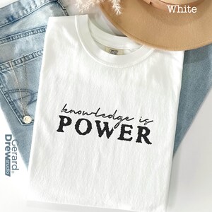 May include: White t-shirt with the text "knowledge is POWER" in black. The shirt is displayed on top of blue jeans, with a tan hat and a silver necklace visible in the background. The shirt is made of a soft material.