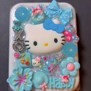 May include: A white rectangular phone case decorated with a 3D Hello Kitty face, blue bows, flowers, and the word "Happy". The case features various blue and pink embellishments, including glitter and small charms.