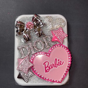 May include: A white rectangular phone case decorated with silver glitter, a silver bow with a pink gem, and the word "DIOR" in silver rhinestones. A pink heart with the word "Barbie" and two star accents complete the design.