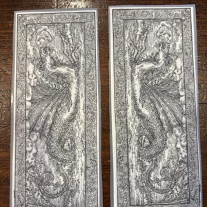 May include: Two rectangular stickers with a detailed black and white illustration of a dragon-like creature with wings and a serpentine body. The design is framed by an ornate border, creating a vintage aesthetic.