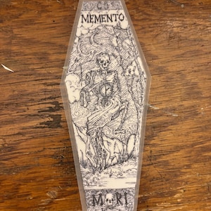 May include: A coffin-shaped sticker with a detailed black and white illustration. The artwork features a skeleton holding a clock, with the words "MEMENTO MORI" at the top and bottom. The sticker has a transparent border.