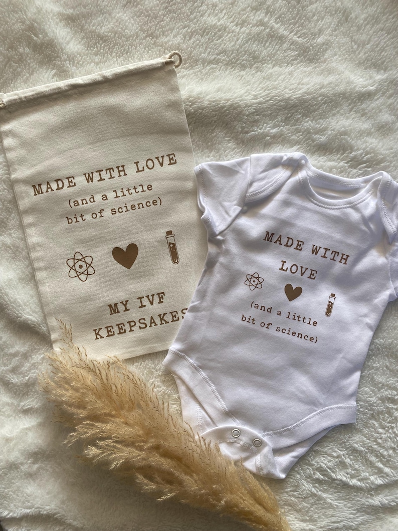 Puede incluir: Una bolsa de lona blanca con el texto "MADE WITH LOVE (and a little bit of science)" y un body blanco con el texto "MADE WITH LOVE (and a little bit of science)". Ambos art&iacute;culos presentan un coraz&oacute;n, un tubo de ensayo y un s&iacute;mbolo de &aacute;tomo.