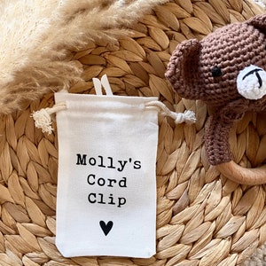 Może przedstawiać: Biała torba ze sznurkiem do ściągania z nadrukiem "Molly's Cord Clip" i czarnym sercem. Torba leży na plecionym słomianym dywaniku.