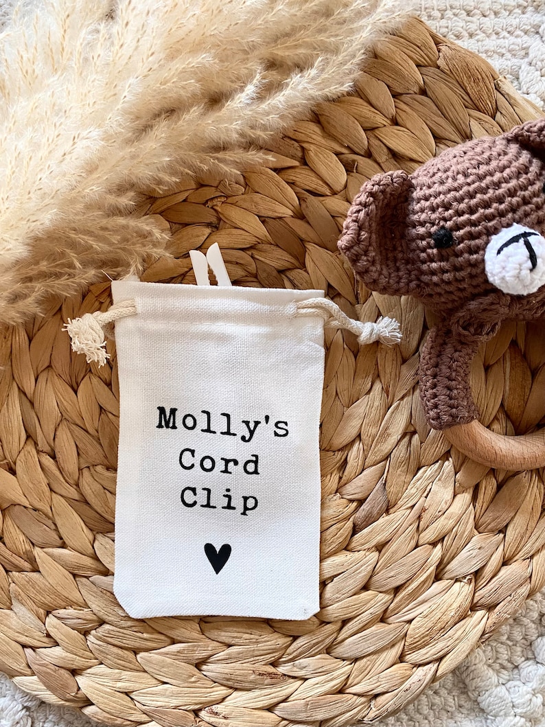 Może przedstawiać: Biała torba ze sznurkiem do ściągania z nadrukiem "Molly's Cord Clip" i czarnym sercem. Torba leży na plecionym słomianym dywaniku.