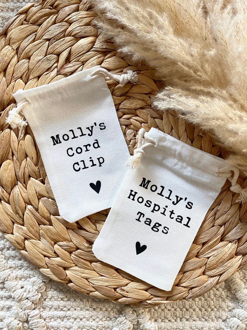 Może przedstawiać: Dwie małe białe materiałowe torebki ze sznurkiem, czarnym tekstem i małym czarnym sercem. Tekst na torebkach brzmi "Molly's Cord Clip" i "Molly's Hospital Tags".