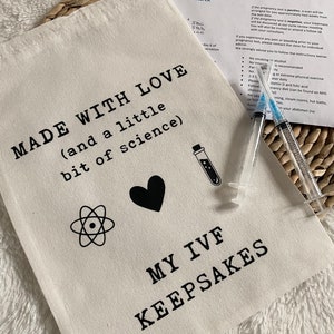 Puede incluir: Una bolsa de lona blanca con el texto "MADE WITH LOVE (and a little bit of science)" y "MY IVF KEEPSAKES". La bolsa tambi&eacute;n presenta un coraz&oacute;n, un s&iacute;mbolo de &aacute;tomo y un tubo de ensayo.