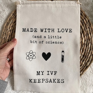 Puede incluir: Una bolsa de lona blanca con el texto "MADE WITH LOVE (and a little bit of science)" y tres iconos: un &aacute;tomo, un coraz&oacute;n y un tubo de ensayo. Debajo del texto se lee "MY IVF KEEPSAKES".