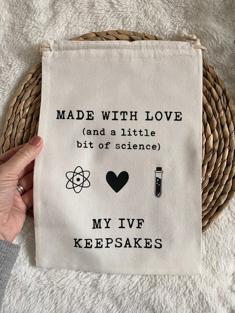 Puede incluir: Una bolsa de lona blanca con el texto "MADE WITH LOVE (and a little bit of science)" y tres iconos: un &aacute;tomo, un coraz&oacute;n y un tubo de ensayo. Debajo del texto se lee "MY IVF KEEPSAKES".