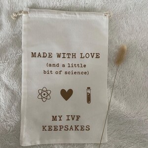 Puede incluir: Una bolsa de tela blanca con el texto "MADE WITH LOVE (and a little bit of science)" y s&iacute;mbolos de un &aacute;tomo, un coraz&oacute;n y un tubo de ensayo. Debajo del texto est&aacute; "MY IVF KEEPSAKES".