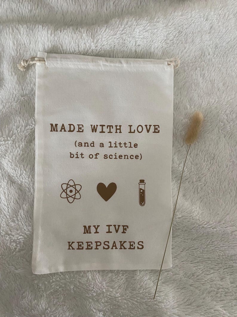 Puede incluir: Una bolsa de tela blanca con el texto "MADE WITH LOVE (and a little bit of science)" y s&iacute;mbolos de un &aacute;tomo, un coraz&oacute;n y un tubo de ensayo. Debajo del texto est&aacute; "MY IVF KEEPSAKES".