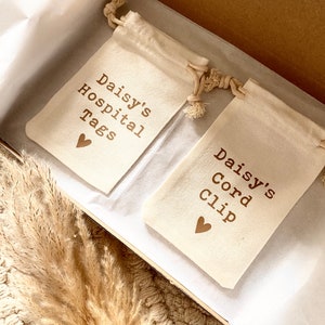 Może przedstawiać: Dwa małe białe worki z sznurkiem i brązowym tekstem. Worki są oznaczone "Daisy's Hospital Tags" i "Daisy's Cord Clip".