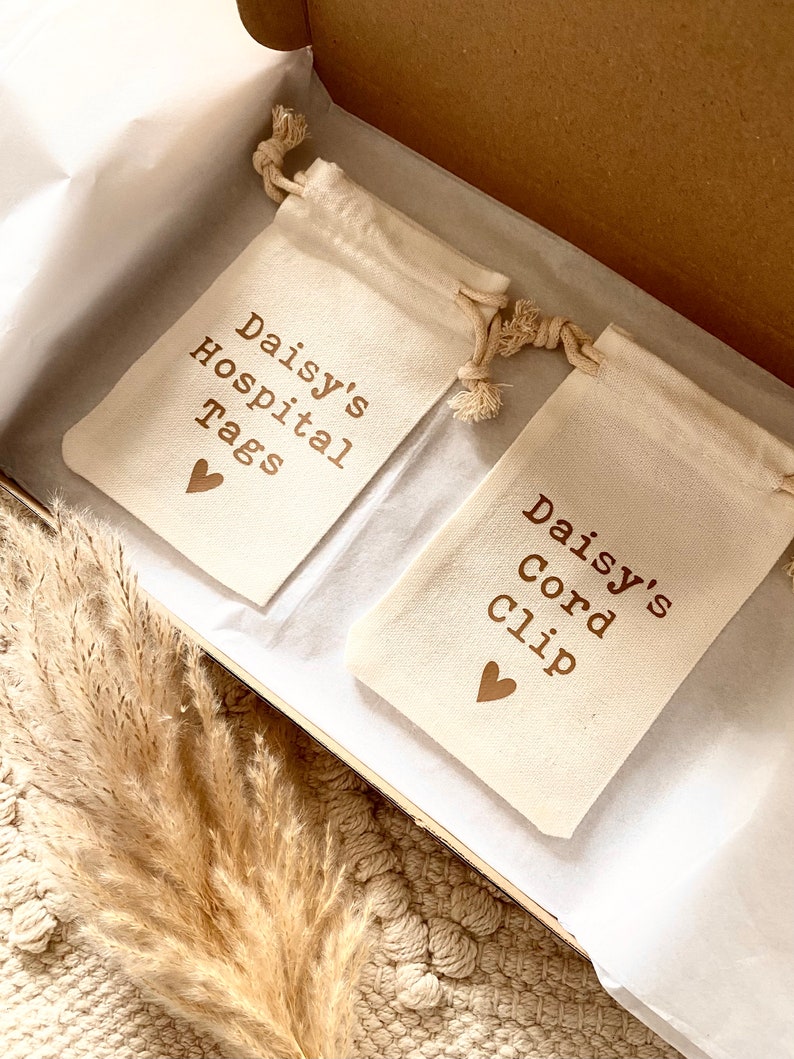 Może przedstawiać: Dwa małe białe worki z sznurkiem i brązowym tekstem. Worki są oznaczone "Daisy's Hospital Tags" i "Daisy's Cord Clip".