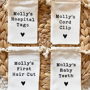 Może przedstawiać: Cztery małe białe materiałowe woreczki ze sznurkiem, z czarnym tekstem i małym czarnym sercem. Woreczki są oznaczone napisami: "Molly's Hospital Tags", "Molly's Cord Clip", "Molly's First Hair Cut" i "Molly's Baby Teeth".