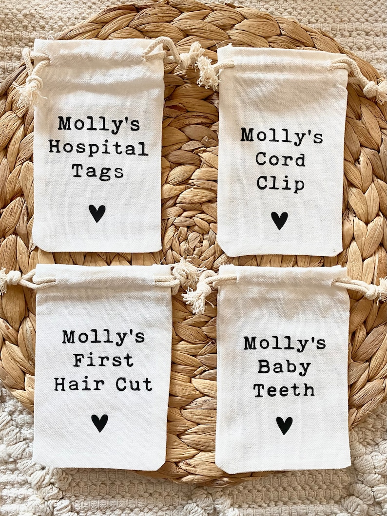 Może przedstawiać: Cztery małe białe materiałowe woreczki ze sznurkiem, z czarnym tekstem i małym czarnym sercem. Woreczki są oznaczone napisami: "Molly's Hospital Tags", "Molly's Cord Clip", "Molly's First Hair Cut" i "Molly's Baby Teeth".