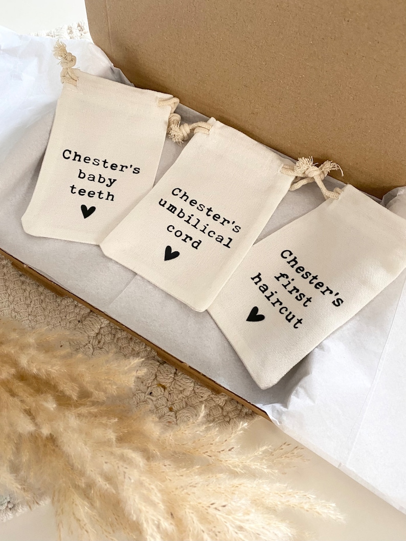Może przedstawiać: Trzy małe białe materiałowe woreczki ze sznurkiem do ściągania i czarnym tekstem. Woreczki są oznaczone napisami "Chester's baby teeth", "Chester's umbilical cord" i "Chester's first haircut". Każdy woreczek ma małe czarne serce na dole.