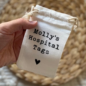 Może przedstawiać: Biały bawełniany woreczek ze sznurkiem do ściągania z czarnym tekstem, kt&oacute;ry brzmi "Molly's Hospital Tags" i małym czarnym sercem.