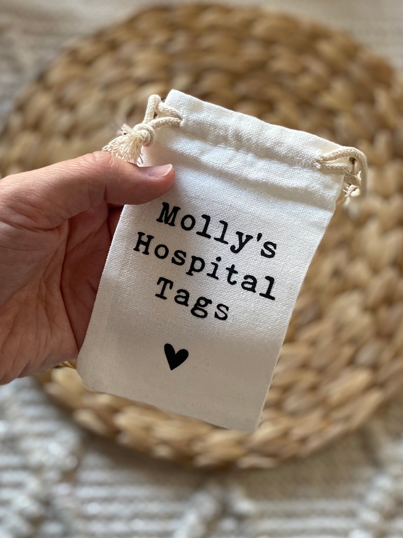 Może przedstawiać: Biały bawełniany woreczek ze sznurkiem do ściągania z czarnym tekstem, kt&oacute;ry brzmi "Molly's Hospital Tags" i małym czarnym sercem.