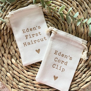Może przedstawiać: Dwie małe białe torby materiałowe z brązowymi sznurkami do zamykania. Torby mają brązowy tekst, kt&oacute;ry brzmi "Eden's First Haircut" i "Eden's Cord Clip". Każda torba ma nadrukowane małe brązowe serce.
