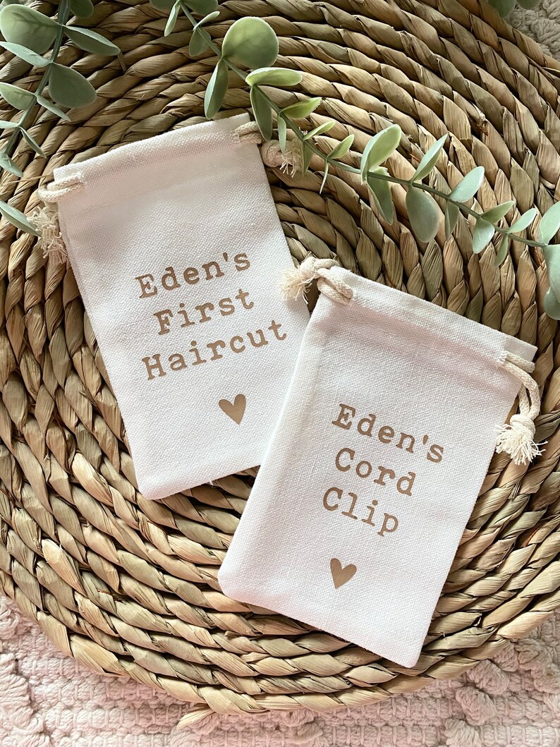Może przedstawiać: Dwie małe białe torby materiałowe z brązowymi sznurkami do zamykania. Torby mają brązowy tekst, kt&oacute;ry brzmi "Eden's First Haircut" i "Eden's Cord Clip". Każda torba ma nadrukowane małe brązowe serce.